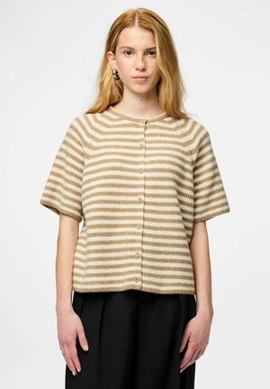 Kvinde med langt blondt hår iført beige og hvide stribede kortærmede knappede cardigan og sorte bukser, set forfra mod en ensfarvet baggrund.