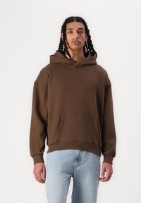 Sweatshirt com capuz castanho em material suave, apresentando um bolso frontal e punhos canelados. Usado com jeans azul claro, o modelo está posicionado contra um fundo branco.