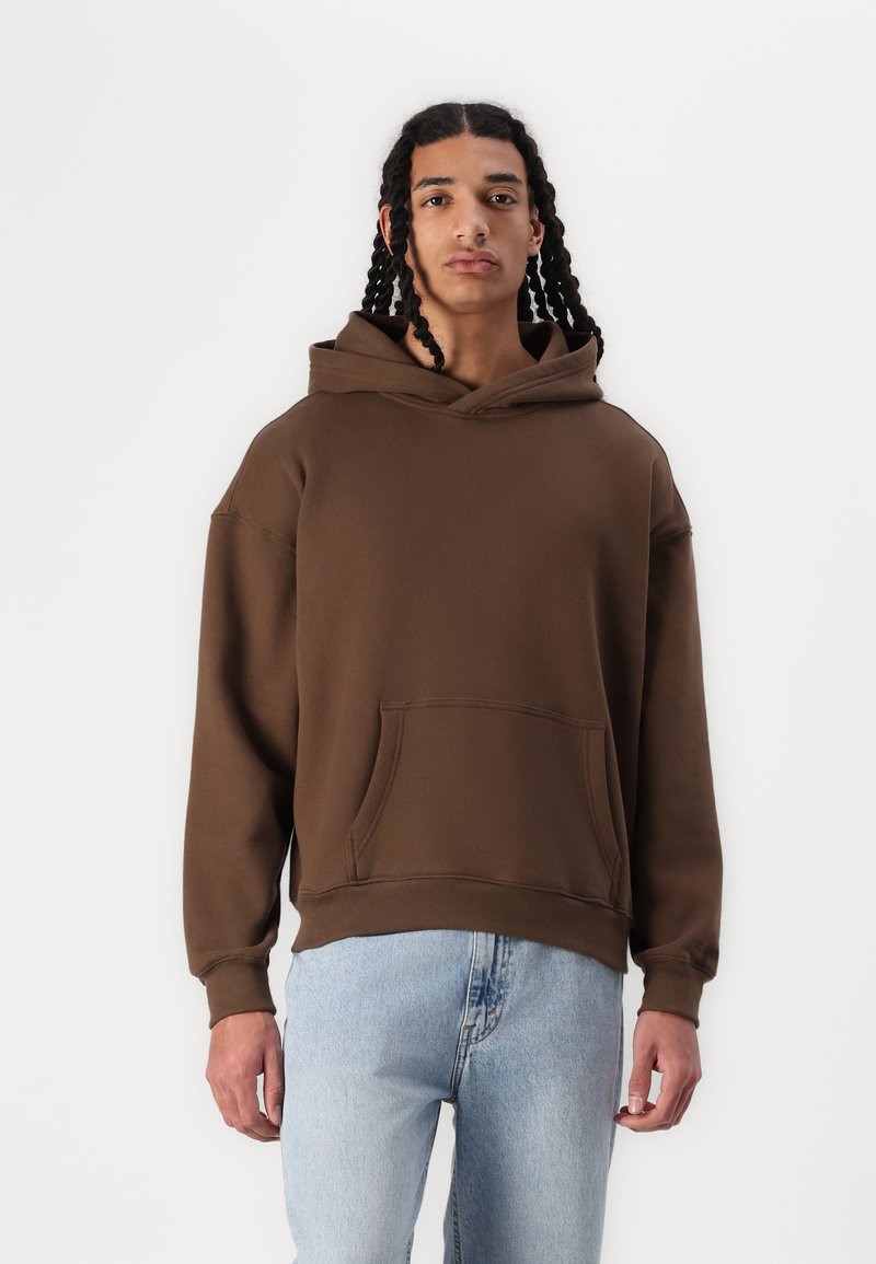 Sweatshirt com capuz castanho em material suave, apresentando um bolso frontal e punhos canelados. Usado com jeans azul claro, o modelo está posicionado contra um fundo branco.