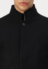 Svart ullkåpe med høy krage, med en enkelt knappelukking og glidelås foran. Glatt tekstur og minimalistisk design.