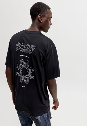 Homme portant un t-shirt noir oversize avec un texte blanc "ATHLETIC PURSUIT" et un design abstrait dans le dos, associé à un short foncé à motifs.