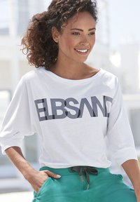 Weißes Langarmshirt mit dem Text "ELBSAND" in fetten schwarzen Buchstaben. Getragen mit grünen Hosen, die einen Kordelzugbund haben.