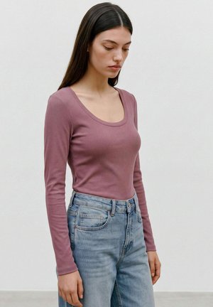 Woman with long dark hair wearing a mauve long-sleeve top and light blue high-waisted jeans standing with her head slightly down.