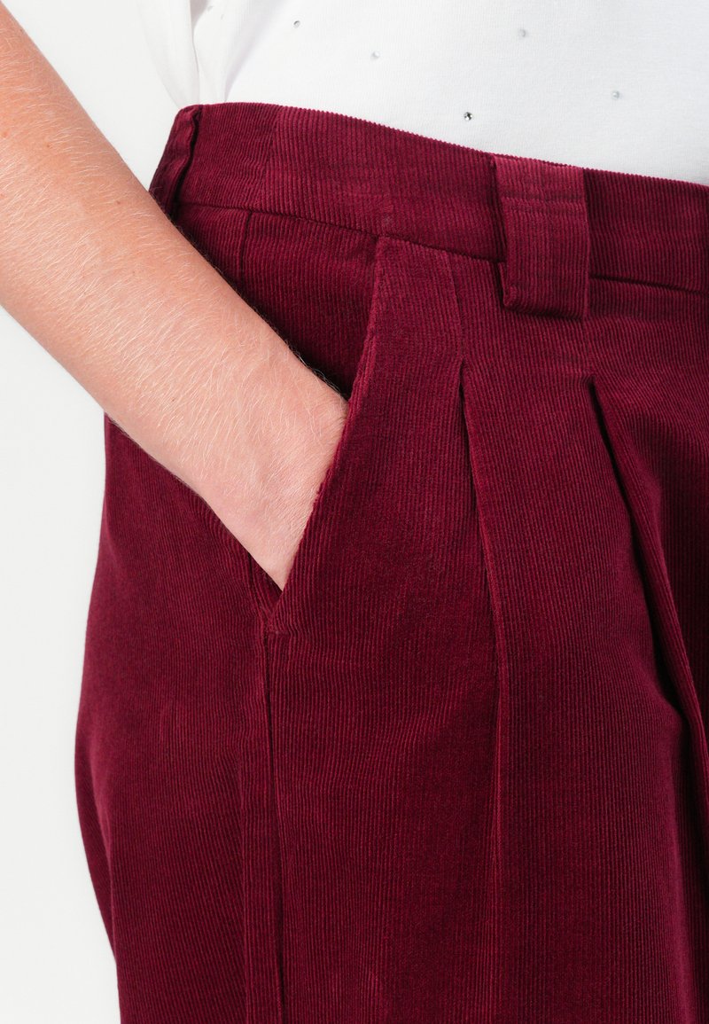 Pantaloni in corduroy bordeaux con tasche laterali, vita alta e texture a righe verticali. Il dettaglio della parte anteriore plissettata aggiunge struttura.