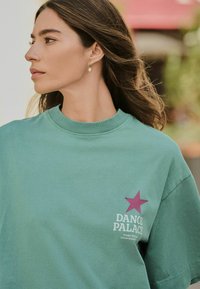 Tricou oversized de culoare verde mentă din bumbac, cu o stea roz și text alb "DANCE PALACE" pe față, având un decolteu amplu.