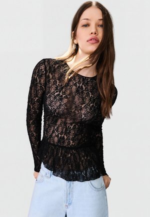 Junge Frau mit langen Haaren, die ein schwarzes, florales Spitzenoberteil mit langen Ärmeln und hellblaue Jeans vor einem schlichten Hintergrund trägt.