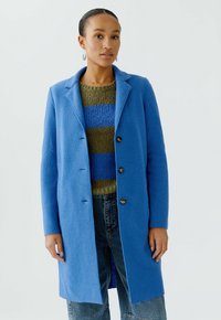 Blauwe wollen mix jas met opstaand revers, knoopsluiting aan de voorkant en twee knopen. Draag het over een groen-blauwe gestreepte trui en denim jeans.