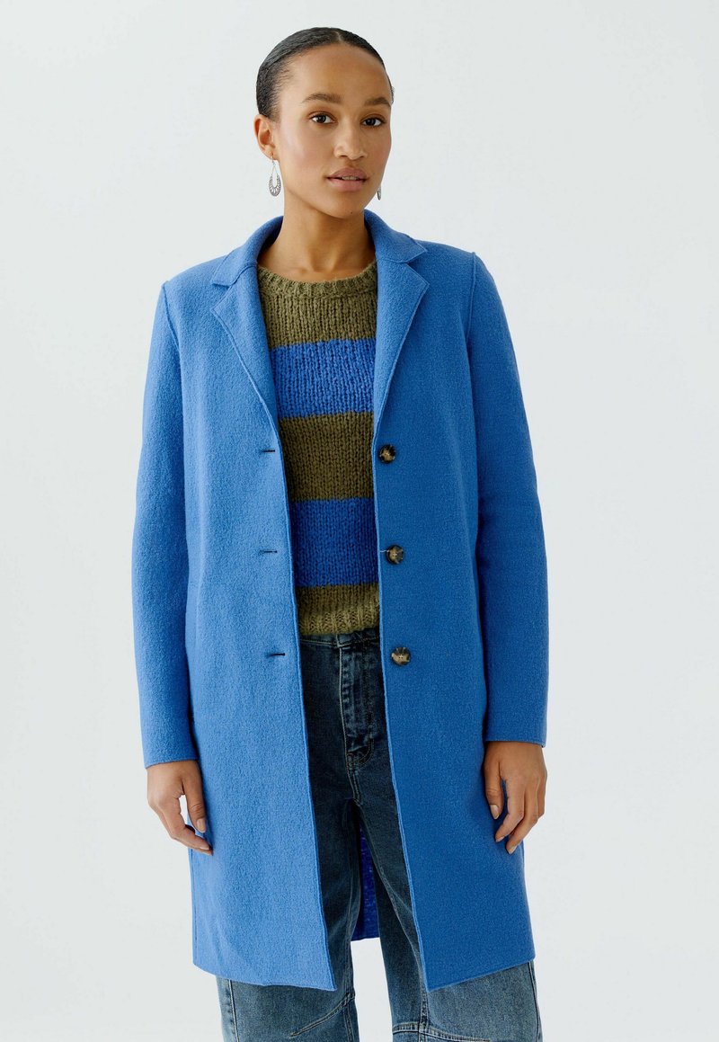 Blauwe wollen mix jas met opstaand revers, knoopsluiting aan de voorkant en twee knopen. Draag het over een groen-blauwe gestreepte trui en denim jeans.