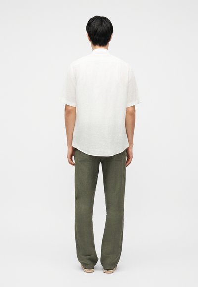 Άνδρας που στέκεται με την πλάτη προς τα έξω, φορώντας λευκό κοντομάνικο πουκάμισο, παντελόνι ελιάς και μπεζ παπούτσια μπροστά σε λευκό αφαιρετικό φόντο.