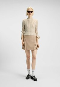 Beige geribbelde langmouwtop met een aansluitend ontwerp en gerimpelde manchetten, gecombineerd met een tan geplisseerde mini-jurk. Zwarte zonnebril en oxford schoenen maken de look compleet.