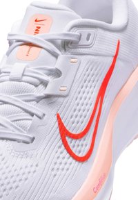 Witte sportieve schoen met een gestructureerde bovenkant, felrode swoosh-logo en een zachte roze zool. Heeft een vetersluiting en gaasdetails voor ventilatie.