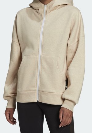 Beige hoodie met rits, kangoeroezak, geribbelde boorden en capuchon, gedragen over een zwarte broek.