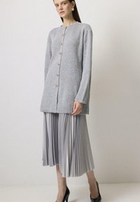 Szary prążkowany kardigan z brązowymi guzikami, zestawiony z plisowaną srebrną spódnicą. Stylizacja charakteryzuje się luźnym krojem oraz długimi rękawami.