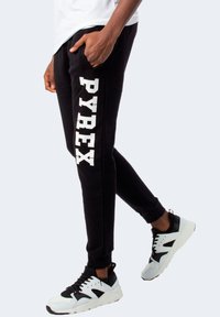 Pantaloni della tuta neri con "PYREX" stampato in grandi lettere bianche lungo il lato, caratterizzati da un design affusolato e polsini a costine, abbinati a scarpe da ginnastica.