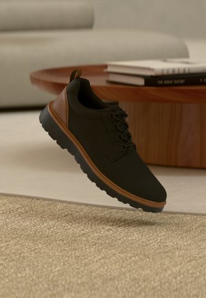 Czarny i brązowy casualowy sneaker z teksturowaną podeszwą, unoszący się nad beżowym dywanem w salonie z drewnianym stolikiem kawowym i ułożonymi na stos książkami.