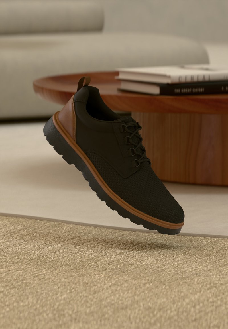 Μαύρο και καφέ αθλητικό παπούτσι με υφή στη σόλα, αιωρούμενο πάνω από μπεζ χαλί σε σαλόνι με ξύλινο τραπεζάκι καφέ και στοιβαγμένα βιβλία.