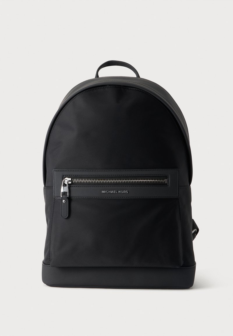 Sac à dos noir Michael Kors avec une poche zippée à l'avant et une poignée supérieure sur un fond clair uni.