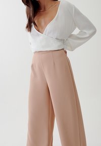 Vitt omslagstopp med djup V-ringning, långa ärmar och textilstruktur, kombinerad med höga, vida ljusrosa byxor.