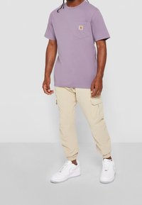 Muž v levandulovém tričku s krátkým rukávem a kapsou na prsou, béžových cargo kalhotách a bílých teniskách na jednobarevném šedém pozadí.