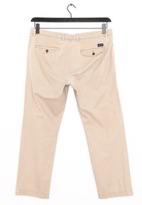 Beige katoenen broek hangend aan een zwarte hanger. Voorzien van twee achterzakken met knoopsluiting en een rechte pijp ontwerp. Gladde textuur.