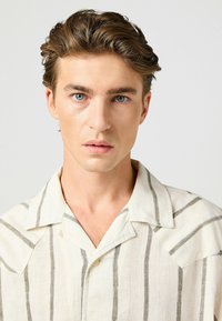 Camicia in lino a maniche corte di colore bianco sporco con strisce verticali grigie; presenta un colletto a punta e chiusura con bottoni nella parte anteriore.