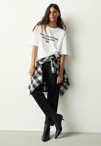 Weißes, übergroßes T-Shirt mit schwarzem Text, schwarze Jeans und ein kariertes Hemd, das um die Taille gebunden ist. Schwarze, spitz zulaufende Stiefeletten runden den Look ab.
