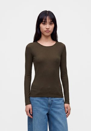 Jeune femme aux longs cheveux foncés portant un haut côtelé à manches longues vert olive et un jean bleu ample, debout devant un fond uni.