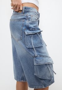 Pantalones cortos de mezclilla tipo cargo en azul claro, con múltiples bolsillos laterales y traseros, dobladillo deshilachado y detalles de costura visibles.