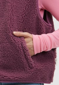 Fleeceweste in tiefem Lila mit strukturierter Oberfläche, seitlichen Taschen und kontrastierendem pinken Langarmshirt darunter. Glatte Innenfutter.