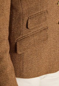 Blazer brown cu model hașură, cu un material texturat țesut, două buzunare cu clapetă și nasturi aurii pentru închidere.