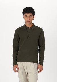 Olive green long-sleeve polo with a zippered collar, made from a soft, textured fabric. Features a fitted silhouette and minimal design.