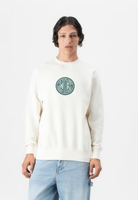 Giovane con capelli ricci corti che indossa una felpa off-white con un logo circolare verde "HUF Worldwide Transit Authority" e jeans azzurri chiari.