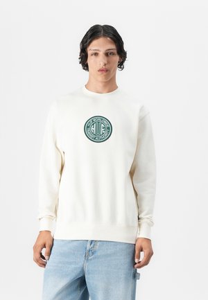 Ung mann med kort krøllete hår iført en off-white sweatshirt med en grønn sirkulær "HUF Worldwide Transit Authority" -logo og lyseblå jeans.