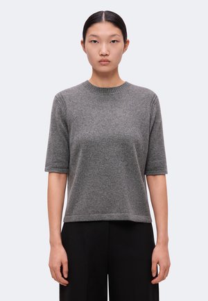 Femme aux cheveux noirs et raides porte un pull tricoté gris à manches courtes et un pantalon noir, debout devant un fond clair uni.