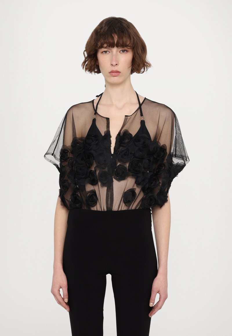 Schwarze transparente Bluse mit floralem Applikationen, die einen tiefen V-Ausschnitt und kurze Ärmel hat. Kombiniert mit enganliegenden schwarzen Hosen.