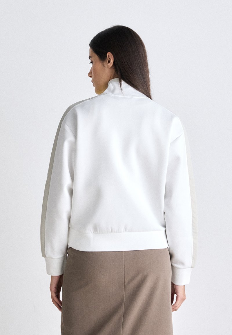 Weißer Pullover mit hohem Kragen, verlängerten Ärmeln mit beigen Akzenten und lockerer Passform. Glatte Textur und gerippte Bündchen.