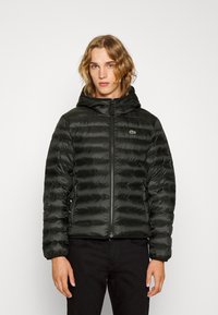 Schwarze, aufgeblasene Jacke mit Kapuze, mit horizontaler Steppung, einem Reißverschluss vorne und einem kleinen grünen Logo auf der linken Brust.