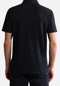 Homme portant un polo noir à manches courtes unies, vu de dos avec les bras détendus sur les côtés, sur un fond uni.