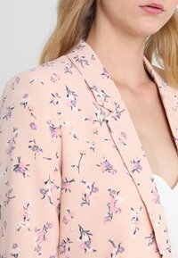 Femme portant un blazer rose clair avec un petit motif floral en nuances de violet, rose et blanc sur un haut blanc.