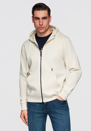 Mężczyzna z zaczesanymi do tyłu włosami, ubrany w kremową bluzę na zamek z kapturem na czarnej koszulce i niebieskich dżinsach, stojący z jedną ręką w kieszeni.