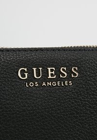 Czarny portfel z teksturowanego tworzywa sztucznego z tłoczonym złotym logo "GUESS" i napisem "LOS ANGELES" poniżej, z zamknięciem na złoty zamek błyskawiczny.