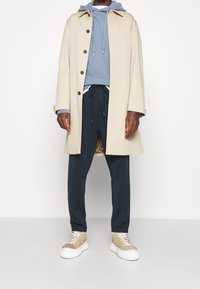 Beige langer Mantel mit braunen Knöpfen über einem blauen Hoodie, kombiniert mit marineblauen Jogginghosen und beigen Sneakers mit weißen Sohlen. Glatte Textur.