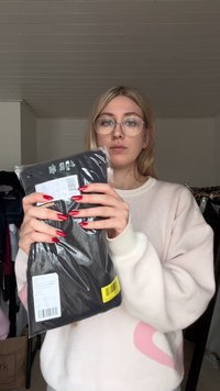 Melnas drēbes gabals caurspīdīgā iepakojumā, kas turēts rokās. Uz iepakojuma redzama baltas krāsas cenu etiķete. Fonā redzami pakarinātāji ar apģērbiem.