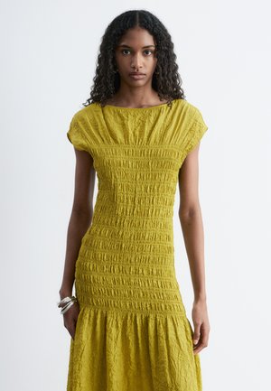 Kobieta z długimi kręconymi włosami, ubrana w teksturowaną żółtą sukienkę z krótkim rękawem i srebrne bransoletki, stoi na tle jednolitego jasnego tła.