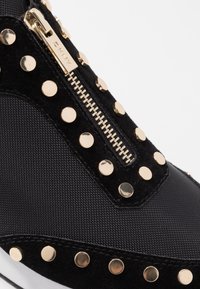 Svarta sneakers med en blandning av mocka och texturerad tyg, guldspikar längs sidan och en detalj med guldblixtlås.