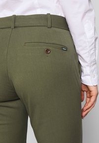 Olivgröna byxor med en slät struktur, med en bakficka med en brun knapp och "Polo"-etikett. Bälteshällor syns vid midjan.