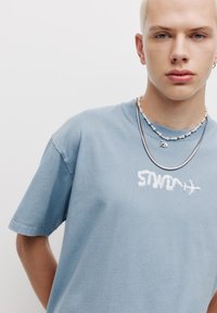 Tricou din bumbac albastru cu text brodat alb, croială lejeră, mâneci scurte, accessorii cu coliere din argint și perle.