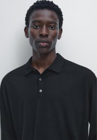 Zwart poloshirt met lange mouwen, zachte textuur, twee knopen aan de hals en een klassieke kraag. Minimalistisch ontwerp, zonder zichtbare patronen of logo's.