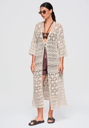 Donna che indossa un lungo cardigan crema lavorato a uncinetto sopra costume da bagno marrone e shorts, con occhiali da sole e sandali neri a ciabatta, in piedi su sfondo semplice.