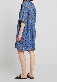 Modrá šatová šaty s oranžovými vzory tygra a palmy, krátkými rukávy a volnějším střihem. Nosené s černými skluznými sandály.